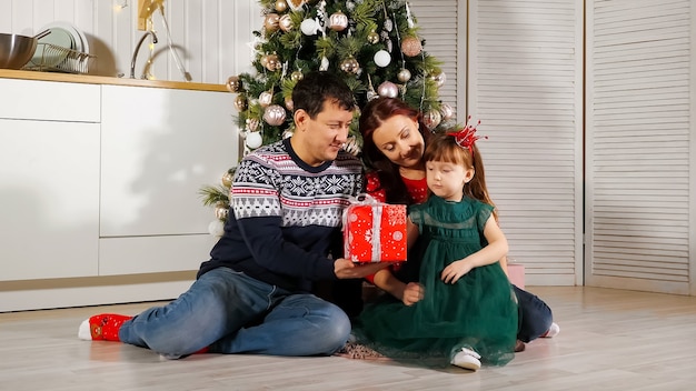 Menina desembrulhando caixa de presente com os pais perto da árvore de natal