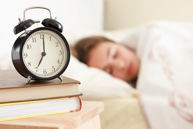 Menina do adolescente que dorme em uma cama branca. Despertador em primeiro plano em uma pilha de livros