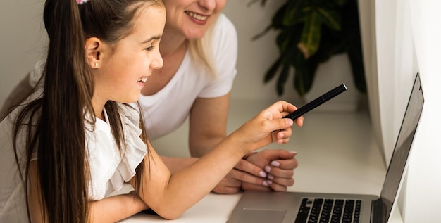 Menina do ensino doméstico aprendendo aula on-line com professor da escola por reunião digital remota na Internet devido à pandemia de coronavírus. Criança procurando computador e escrevendo nota com a ajuda da mãe.