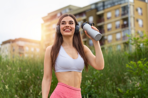 Menina do esporte urbano com água potável de fones de ouvido no pôr do sol.