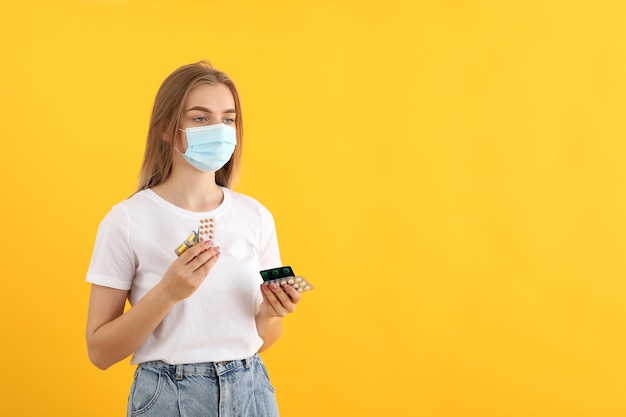 Menina doente com máscara e comprimidos em fundo amarelo