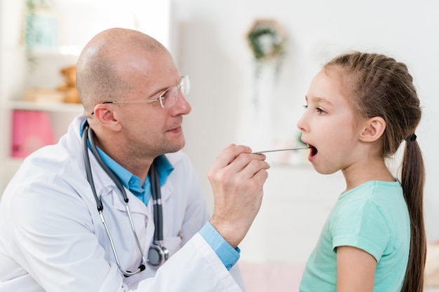 Menina doente mantendo a boca aberta enquanto o médico de jaleco examina sua garganta inflamada com um instrumento médico