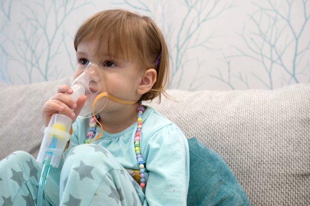 Menina doente segurando o inalador na mão e respirando através de um nebulizador em casa