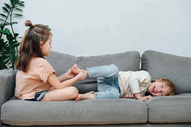 Menina dominante fazendo cócegas para fazer cócegas no pé dos meninos, fazendo-o rolar e rir na vista lateral do sofá