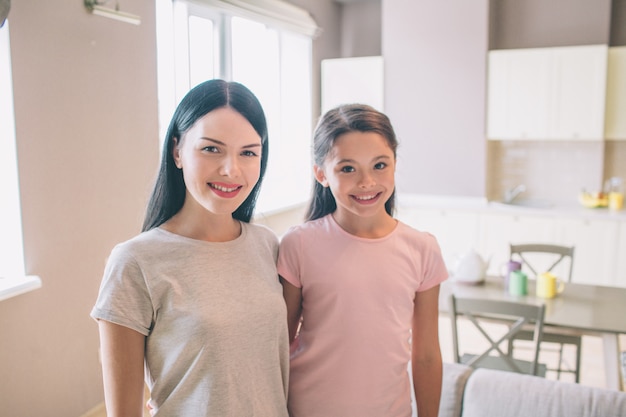 Menina e a mãe dela estão de pé na cozinha e posando na câmera. eles sorriem. boas damas estão se abraçando.