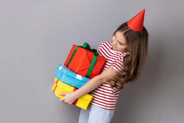 Menina e no cone de festa segurando uma pilha pesada de presentes comemorando seu aniversário