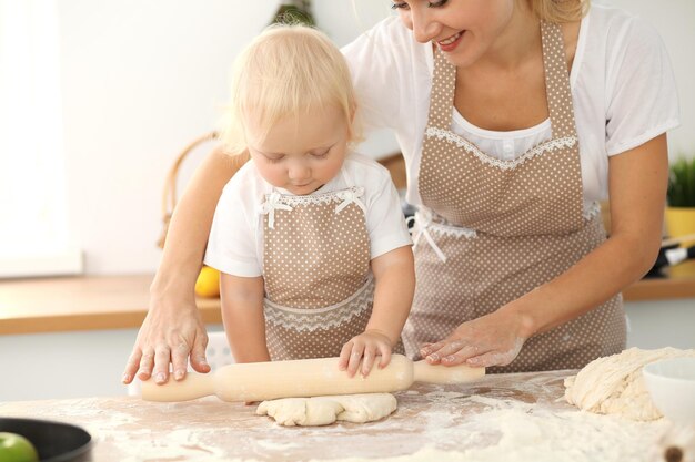 Menina e sua mãe loira em aventais bege brincando e rindo enquanto amassa a massa na cozinha. Pastelaria caseira para pão, pizza ou biscoitos. Diversão em família e conceito de culinária.