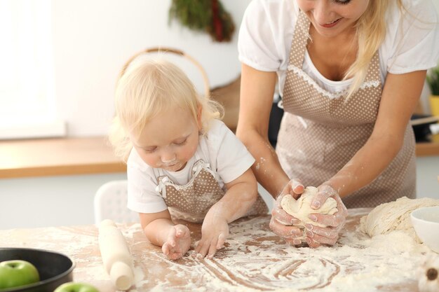 Menina e sua mãe loira em aventais bege brincando e rindo enquanto amassa a massa na cozinha. Pastelaria caseira para pão, pizza ou biscoitos. Diversão em família e conceito de culinária.