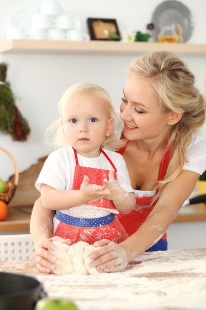 Menina e sua mãe loira em aventais vermelhos brincando e rindo enquanto amassava a massa na cozinha. Pastelaria caseira para pão, pizza ou biscoitos. Diversão em família e conceito de culinária.