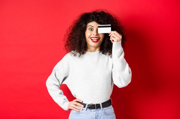 Menina elegante com cabelo encaracolado, mostrando o cartão de crédito de plástico sobre o olho e sorrindo, de pé contra a parede vermelha.