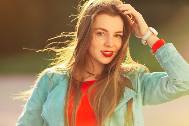 Menina elegante com lábios vermelhos mantém o cabelo em luz suave ao pôr do sol. Retrato de mulher jovem, usando um relógio, blazer ciano brilhante e blusa vermelha Olha para a câmera