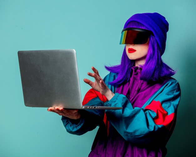 Menina elegante com óculos cyber punk e agasalho segurando um laptop na parede azul