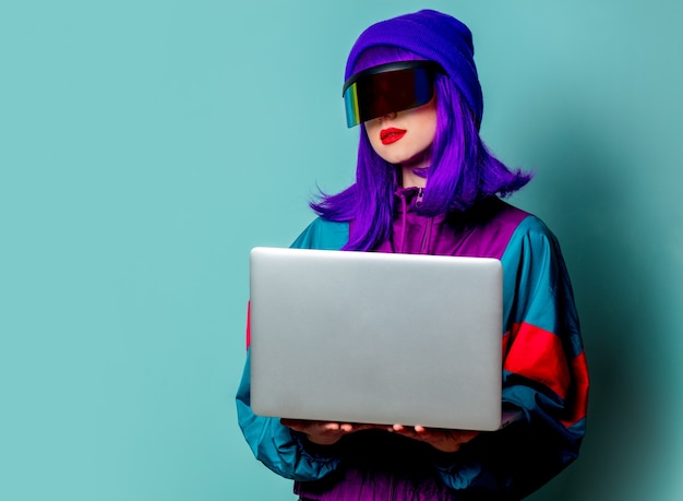 Menina elegante com óculos cyber punk e agasalho segurando um laptop na parede azul