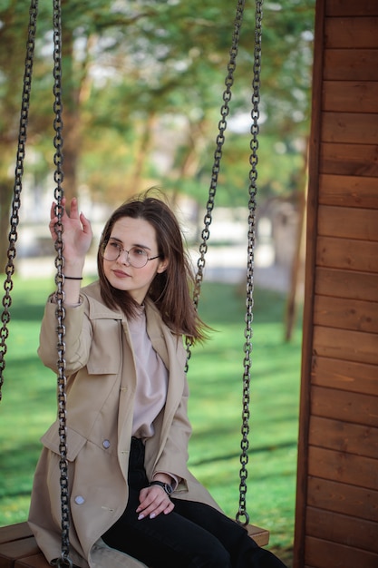 Menina elegante com óculos parece longe, ao ar livre. Menina adolescente em um balanço, retrato bonito.