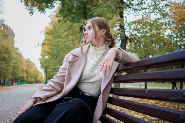 Menina elegante com um casaco se senta em um banco e olha para a rua