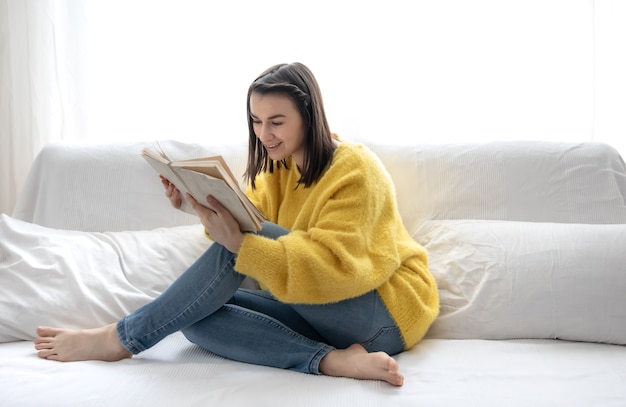 Menina elegante com um suéter amarelo está lendo um livro com entusiasmo enquanto está sentado no sofá em casa.