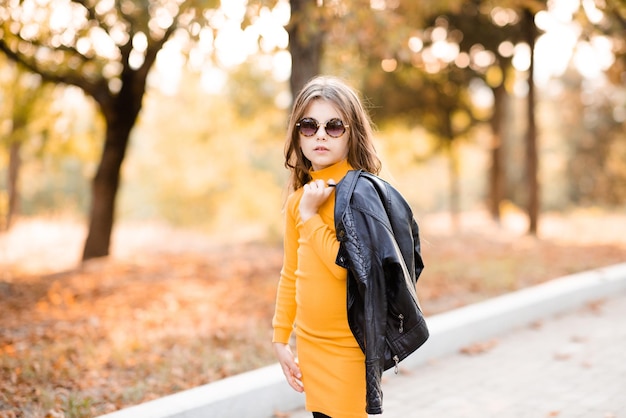 Menina elegante de 4 a 5 anos usa vestido de malha amarelo e óculos de sol seguram jaqueta de couro preta