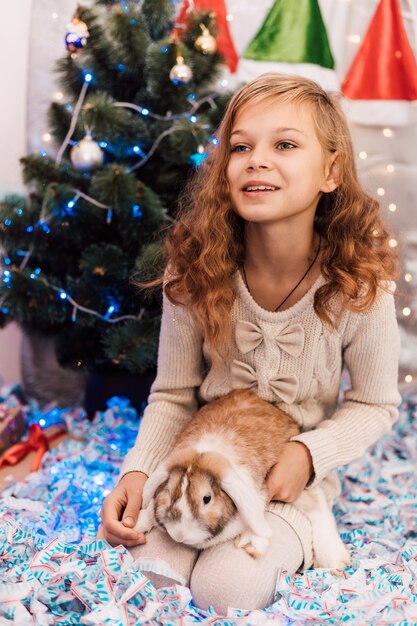 Menina elegante de ano novo em um vestido leve perto de um abeto decorado de verde segurando um coelho