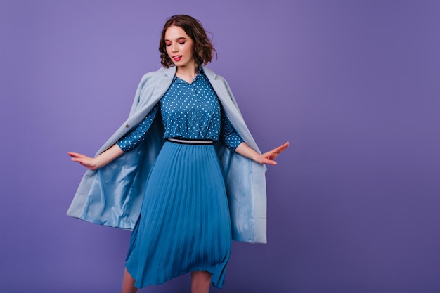 Menina elegante de cabelos escuros dançando em fundo roxo Foto interior de mulher feliz com penteado curto usa casaco azul
