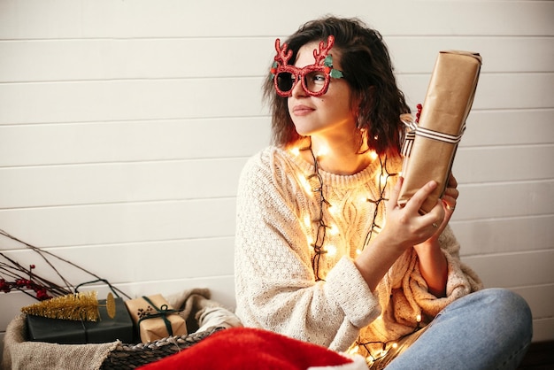 Foto menina elegante e feliz em copos festivos com chifres de rena agitando o presente de natal e sorrindo nas luzes de natal mulher jovem hipster em suéter aconchegante com presente de natal boas festas