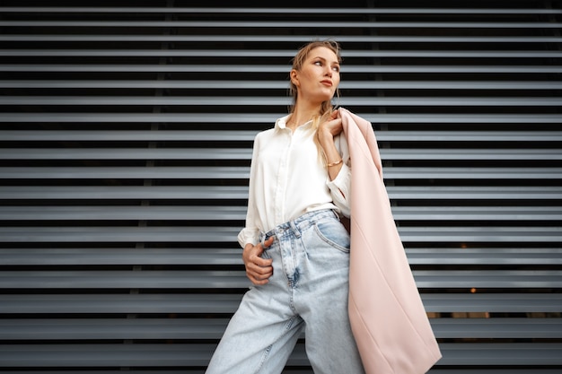 Menina elegante em jeans e blusa de algodão branca posando perto de uma parede cinza