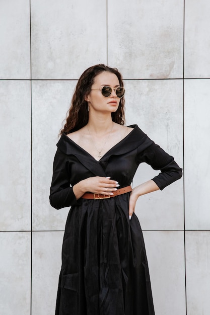 Menina elegante em um vestido preto e óculos de sol perto da parede cinza. Foto de mulher de óculos. Mulher jovem, penteado encaracolado, sorrindo perto de uma parede na rua. Garota vestindo roupas de vestido preto.