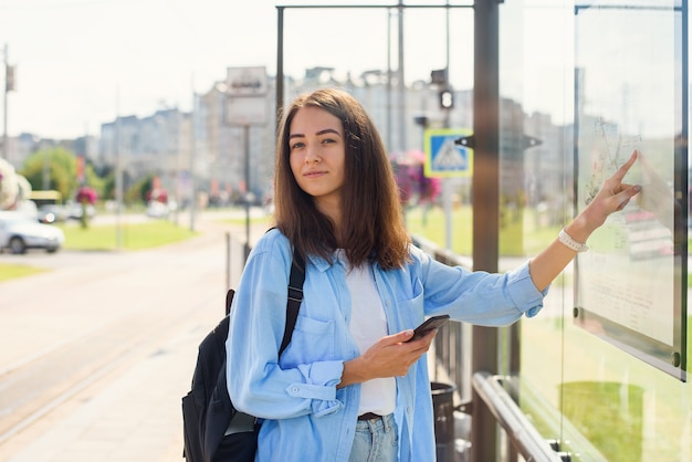 Menina elegante olha para o mapa de transportes públicos para encontrar a estação certa em pé na moderna estação de bonde ao ar livre