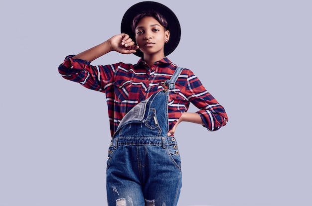 Menina elegante preto hipster com cabelo curto em uma camisa xadrez e macacão jeans.