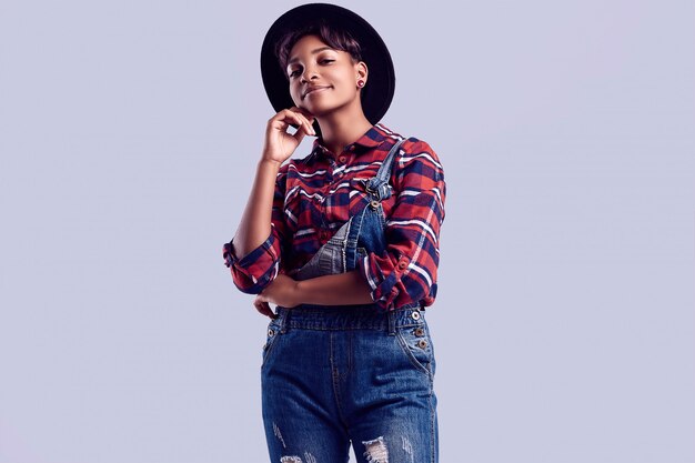 Menina elegante preto hipster com cabelo curto em uma camisa xadrez e macacão jeans.