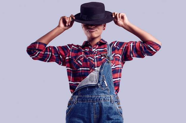 Menina elegante preto hipster com cabelo curto em uma camisa xadrez e macacão jeans.