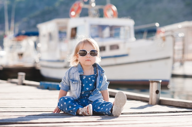 Menina elegante vestindo roupas jeans da moda sentada no cais de madeira ao ar livre