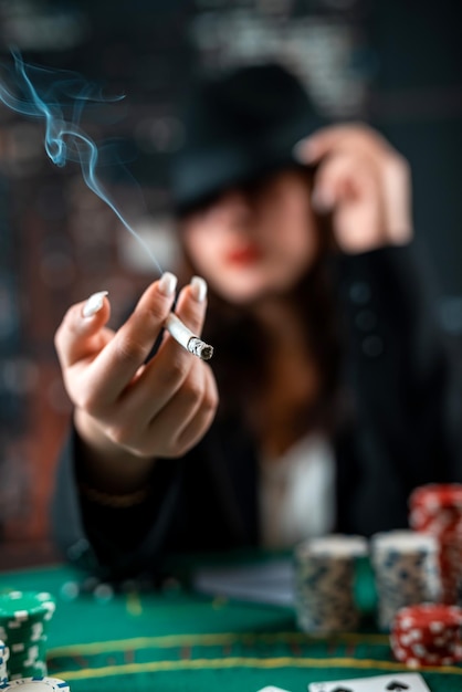 Menina elegantemente vestida fuma um cigarro sopra fumaça e joga cartas em uma mesa sobre um pano verde