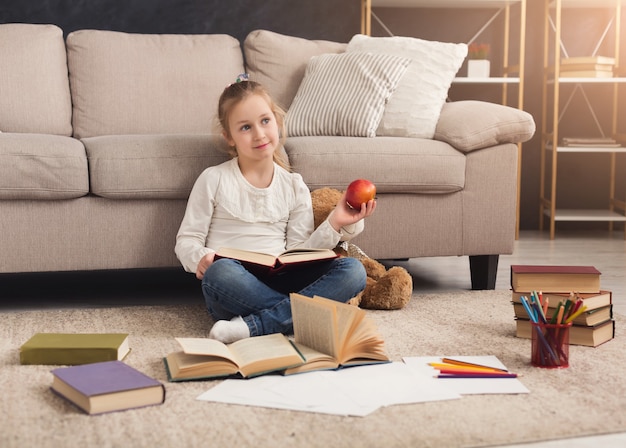 Menina em casa com o livro sentado no tapete e comendo maçã. Criança lendo histórias emocionantes, sentada perto do ursinho de pelúcia. Divertido e fácil conceito de educação lúdica, copie o espaço