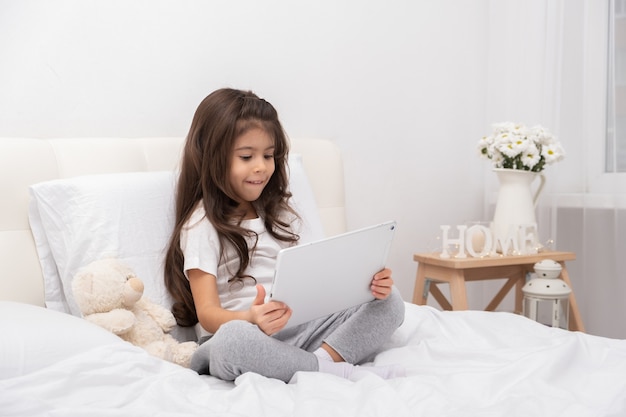 Menina em casa com o ursinho de pelúcia usando o tablet sentado na cama.