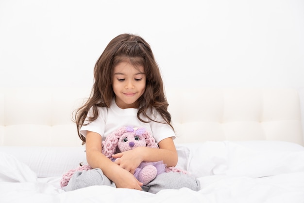 Menina em casa vestir se senta com o coelhinho de pelúcia na cama em casa.