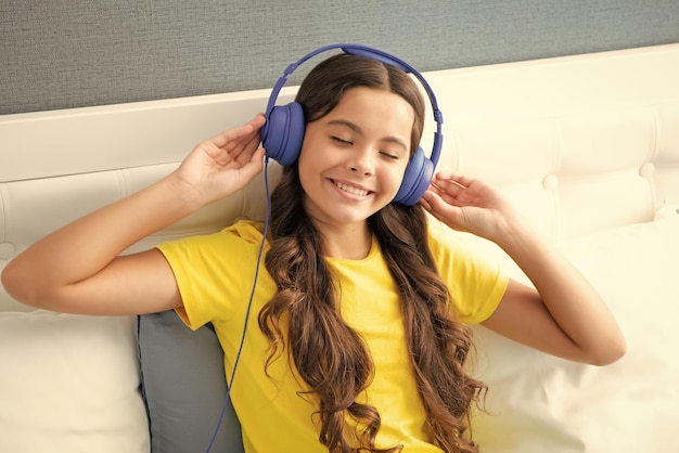 Menina em fones de ouvido relaxa na cama em casa usando fones de ouvido Menina adolescente feliz emoções positivas e sorridentes