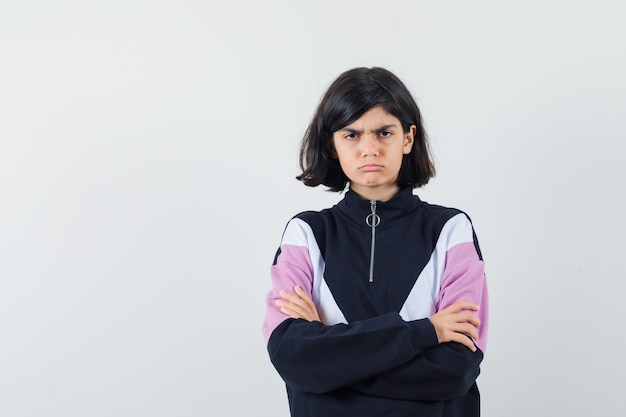 Menina em pé com os braços cruzados na camisa e olhando séria, vista frontal.