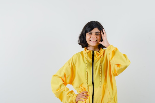 Menina em pé na pose de pensamento com capuz amarelo e olhando alegre, vista frontal.