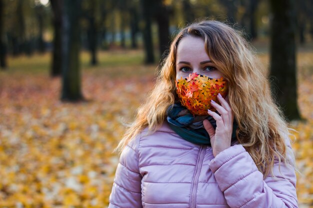 Menina em um fundo de árvores em um parque em uma máscara de um vírus com folhas de outono