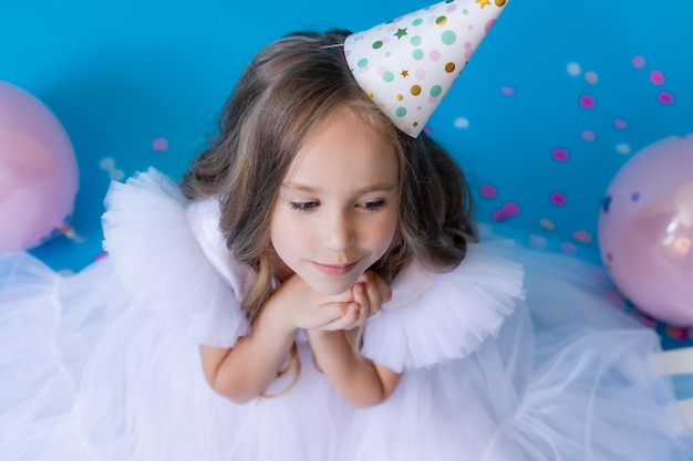 Menina em um lindo vestido branco fofo e um chapéu festivo está sentado no chão sobre fundo azul