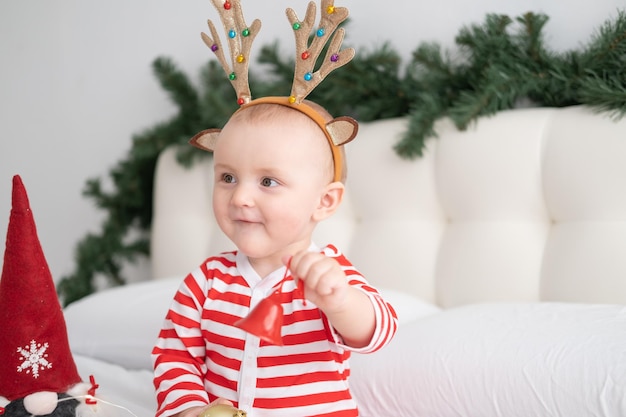 Menina em um macacão listrado com chifres de veado na cabeça brincando com o sino de natal na cama decorada