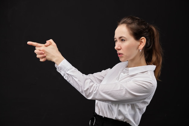 Menina em um sumário branco da camisa que aponta uma arma fora das mãos em um fundo preto.