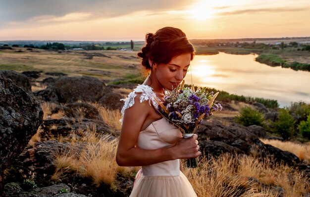 menina em um vestido de noiva cheirando um buquê de casamento ao pôr do sol