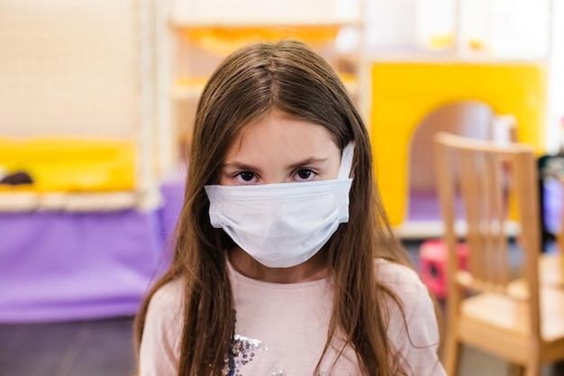 Menina em uma máscara médica interna no retrato de closeup de grupo de brincadeiras em fundo desfocado Uma criança mascarada com olhos escuros e tristes Medidores de segurança pandêmicos afetam a vida das crianças