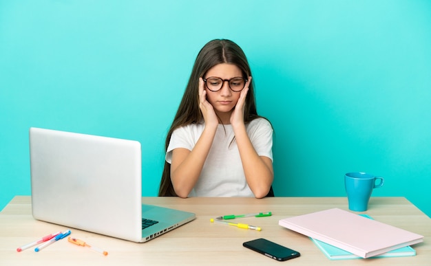 Menina em uma mesa com um laptop na parede azul isolada com dor de cabeça