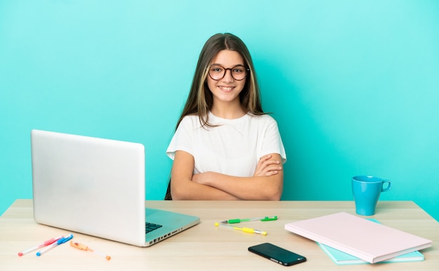 Menina em uma mesa com um laptop sobre fundo azul isolado, mantendo os braços cruzados na posição frontal