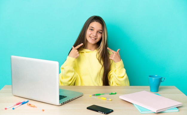 Menina em uma mesa com um laptop sobre um fundo azul isolado fazendo um gesto de polegar para cima