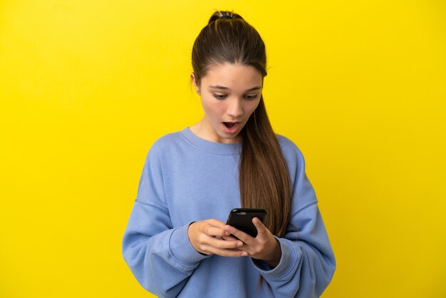 Menina em uma parede amarela isolada olhando para a câmera enquanto usa o celular com expressão de surpresa