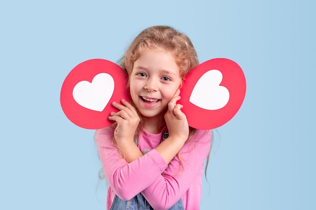 Foto menina encantada com roupas casuais, mantendo ícones de coração perto do rosto e sorrindo amigavelmente enquanto representa as mídias sociais para crianças