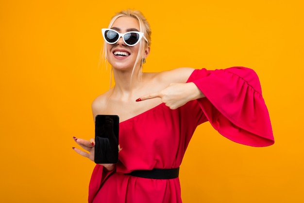 Menina encantadora caucasiana em um vestido vermelho mostra uma tela de telefone em branco em uma parede amarela